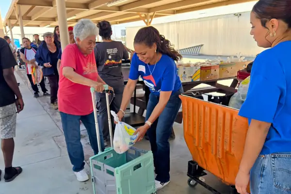 Grocery Distribution webp A volunteer hands a bag of food to an elderly woman using a walker, while others wait in line at a community food distribution event.