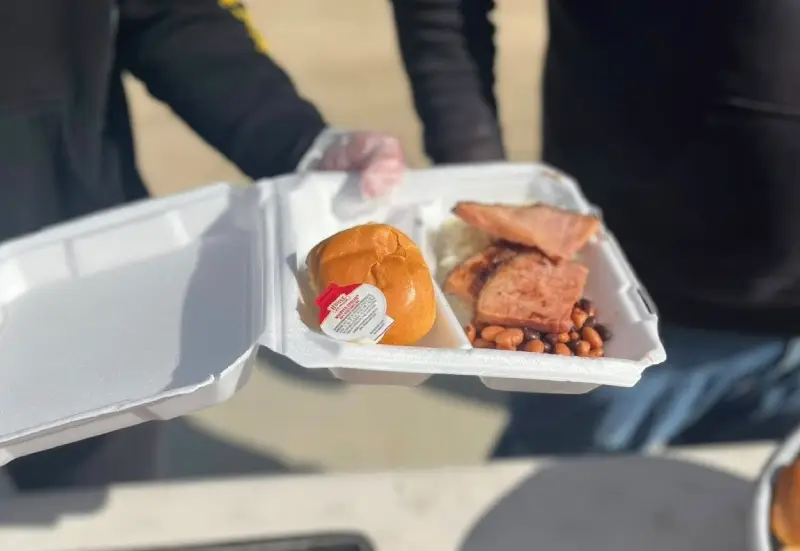 Hotmeals-scaled-e1749673498172-800x551 new webp A person holding a takeout container with a sandwich, meat, baked beans, and sauce in a sunny outdoor setting.
